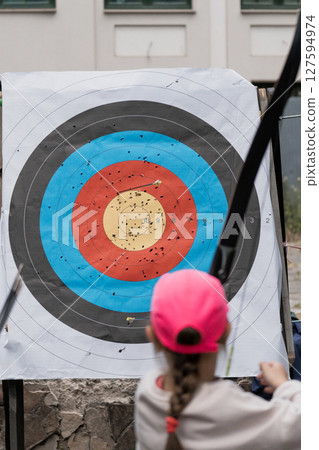 Archery Practice Target with Scattered Arrows and Young Archer in Pink Cap, Outdoor Sport Activity, Blue and Yellow Target, Focus on Precision. Archery Practice Target with Scattered Arrows and Young Archer in Pink Cap, Outdoor Sport Activity, Blue and Yellow Target, Focus on Precision. 127594974