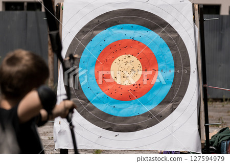Archery Practice at Outdoor Range: Boy Shooting Arrows at Bullseye Target with Scattered Impacts, Wooden Stand, Blue and Brown Colors 127594979