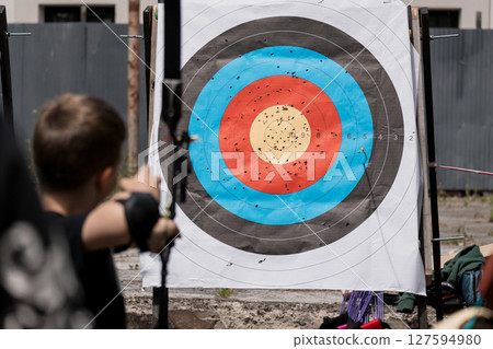 Boy Archery Practice Outdoors: Target Board, Arrows, Backyard, Summer Sports Activity, Blue, Red, Black, Training, Skill Development, Competition. Boy Archery Practice Outdoors: Target Board, Arrows, Backyard, Summer Sports Activity, Blue, Red, Black, Training, Skill Development, Competition. 127594980