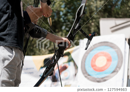 Archery Practice Outdoors: Young Athlete Pulls Back Bowstring, Aiming at Bullseye Target, Focus on Equipment and Nature Background 127594983