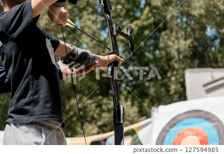 Young Archer Drawing Back Compound Bow Aiming at Target Outdoors, Summer Archery Training, Dark Blue T-Shirt, Green Trees, Sports Lifestyle. 127594985