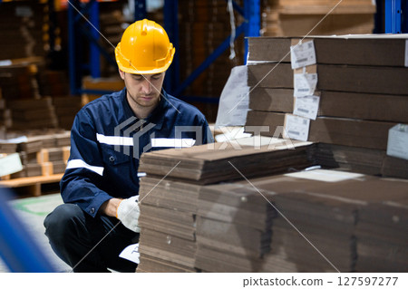 Man paper warehouse worker using clipboard checking inspecting quality cardboard in storage. Large industry in paper product line manufacturing. Employee level wear uniform and helmet yellow. 127597277