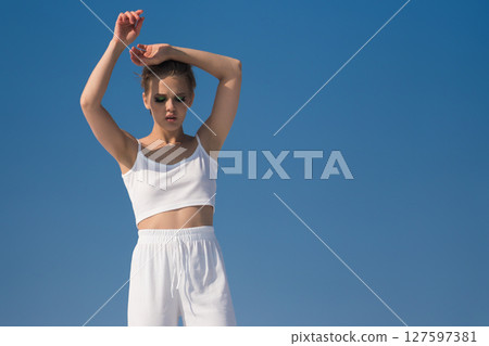 Beautiful female fashion model with her hands raised above her head and her head looking down. Serene sexy woman dressed in white crop top posing modestly against blue sky in sunny summer weather 127597381