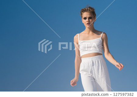 Beautiful fashion model moves against azure sky on sun kissed summer day. Front view sensual Caucasian woman is absolutely captivating in white crop top and pants that accentuate every curve Beautiful fashion model moves against azure sky on sun kissed summer day. Front view sensual Caucasian woman is absolutely captivating in white crop top and pants that accentuate every curve 127597382