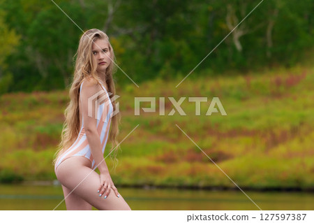 Young slim fashion model with long blonde hair is showcased in one piece swimsuit, standing majestically surrounded by breathtaking autumn forest. Woman from Gen Z enjoying beauty of nature 127597387