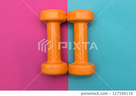 Two orange dumbbells on a pink and blue background. A tool for training, sport and activity in summer time. 127598119