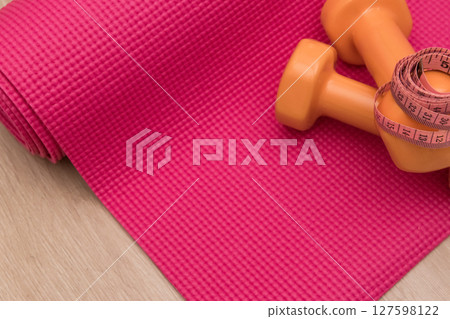 Two orange dumbbells and roulette on a pink yoga mat in fitness studie. A tools for training, sport and activity in summer time. Two orange dumbbells and roulette on a pink yoga mat in fitness studie. A tools for training, sport and activity in summer time. 127598122