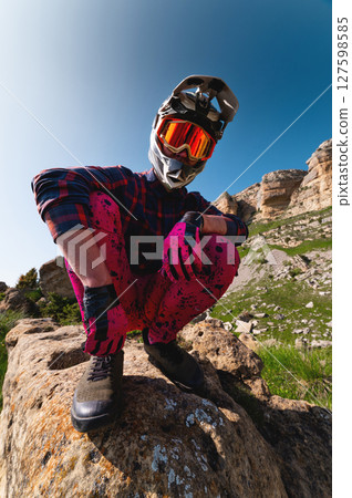 Close-up portrait of young sportsman wearing protective helmet and protective mask on his face. Mature guy resting looking at camera after hard training in mountains Close-up portrait of young sportsman wearing protective helmet and protective mask on his face. Mature guy resting looking at camera after hard training in mountains 127598585