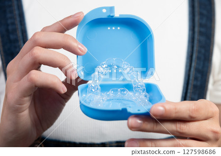 Woman holding clear retainers with box in hands, front view on background of girl's body in overalls and white t-shirt, close-up 127598686