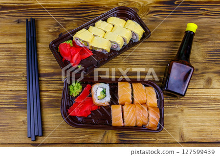 Set of sushi rolls in plastic boxes, soy sauce and chopsticks on wooden table. Top view. Sushi for take away or delivery of sushi in plastic containers 127599439