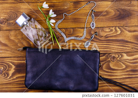 Women accessories on wooden background. Clutch bag, bottle of perfume, necklace, earrings and snowdrops on wood table. Beauty and fashion composition. Top view, flat lay Women accessories on wooden background. Clutch bag, bottle of perfume, necklace, earrings and snowdrops on wood table. Beauty and fashion composition. Top view, flat lay 127601031