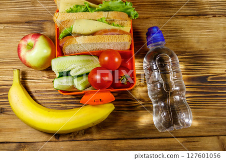 Bottle of water, apple, banana and lunch box with sandwiches and fresh vegetables on a wooden table. Top view 127601056