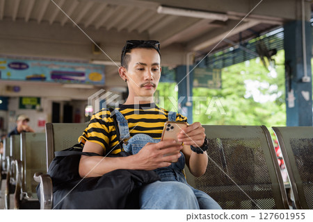 Portrait of nonbinary genderfluid young Asian gay tourist man in bus terminal using phone 127601955