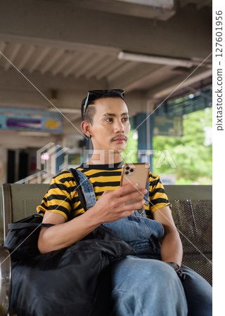 Portrait of nonbinary genderfluid young Asian gay tourist man in bus terminal using phone 127601956