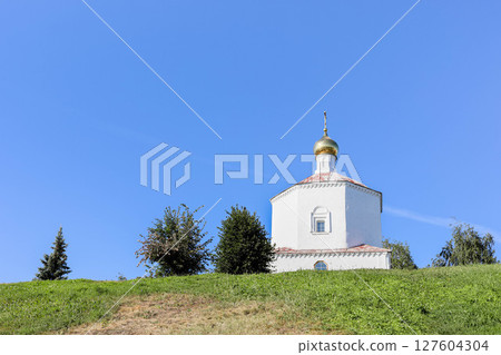Historic white chapel with golden dome on grassy hill under clear blue sky 127604304