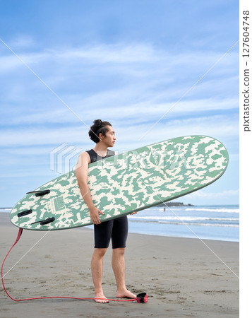 A young man holding a surfboard and quietly gazing at the ocean 127604748