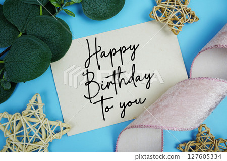 Happy Birthday to you text on paper card with fabric ribbon and rattan star decorations on blue background 127605334
