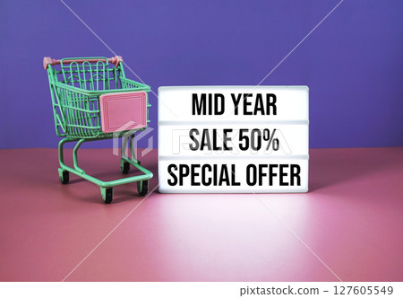 Mid Year Sale 50% off Specials Offer text on LED lightbox with trolley shopping cart on pink and purple geometric background 127605549
