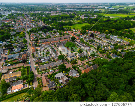 Aerial view of a peaceful suburban neighborhood surrounded by lush greenery, residential houses, and tree-lined streets under a cloudy sky, with fields and water in the distant background. Aerial view of a peaceful suburban neighborhood surrounded by lush greenery, residential houses, and tree-lined streets under a cloudy sky, with fields and water in the distant background. 127606774