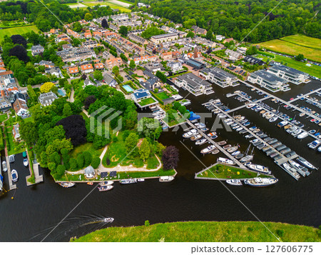 A top-down view of a marina with multiple boats docked along wooden piers, a boat cruising through a narrow waterway surrounded by green parks and natural grassy areas. A top-down view of a marina with multiple boats docked along wooden piers, a boat cruising through a narrow waterway surrounded by green parks and natural grassy areas. 127606775