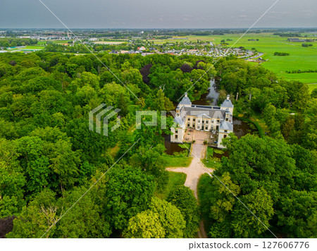 Aerial view of a historic castle surrounded by a forest, with vast green fields, winding waterways, and a river stretching across the flat Dutch countryside under a partly cloudy sky. Aerial view of a historic castle surrounded by a forest, with vast green fields, winding waterways, and a river stretching across the flat Dutch countryside under a partly cloudy sky. 127606776