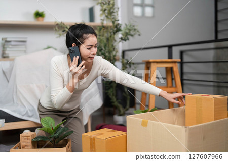 Moving and Communication. Young woman talking on phone while organizing boxes for relocation. 127607966