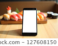 A black cell phone is sitting on a wooden table with a plate of sushi 127608150