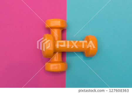 Two orange dumbbells on a pink and blue background. A tool for training, sport and activity in summer time. 127608363