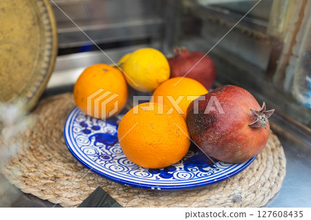 Citrus and pomegranate fruits rest on a traditional blue patterned plate. Mediterranean freshness, market display, and rustic natural nutrition. Citrus and pomegranate fruits rest on a traditional blue patterned plate. Mediterranean freshness, market display, and rustic natural nutrition. 127608435