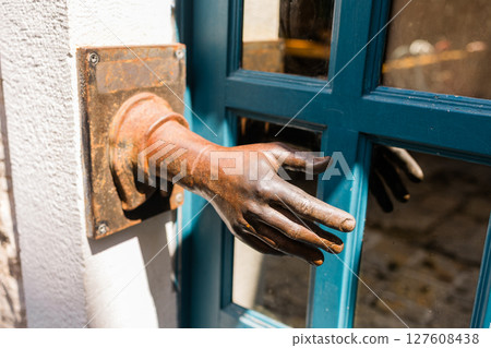 Rusty sculpted hand emerges from a wall beside a blue wooden door. Artistic urban decor, surreal visual element, and unique street detail in Budva, Montenegro. Rusty sculpted hand emerges from a wall beside a blue wooden door. Artistic urban decor, surreal visual element, and unique street detail in Budva, Montenegro. 127608438