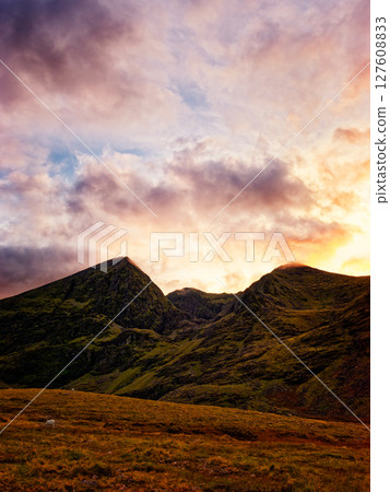 Serene sunset paints the rugged peaks of the Highlands in vibrant hues, casting a warm glow on the tranquil valley below. A solitary sheep grazes peacefully. 127608833
