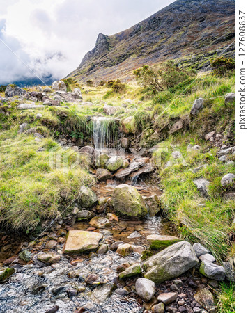 A small waterfall cascades over rocks and vegetation in a picturesque landscape. This is along the popular hiking trail of Carrauntoohill in County Kerry, Ireland, with a mountain in the background. 127608837