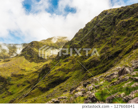 Hills covered in green vegetation and scattered rocks form an Irish mountain landscape. Mist and clouds roll through the valleys near Carrauntoohil along the hiking trail in County Kerry. 127608841