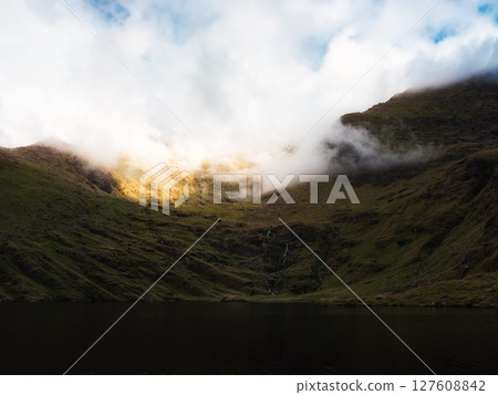 A still body of water is nestled between rolling, grass covered hills that rise into a mountain partially shrouded in white fog near Lough Callee in the Irish countryside. 127608842