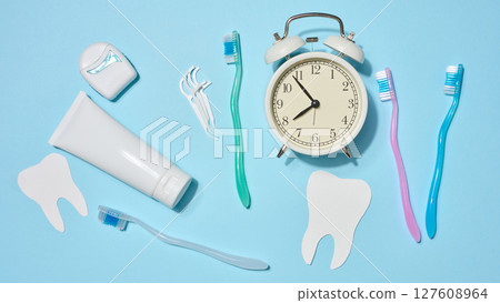 White tube of toothpaste, dental floss in a case, toothbrushes of different colors, white alarm clock, plastic flossers placed on a blue background. Top view. 127608964