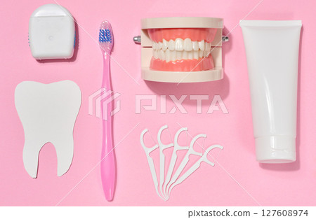 White tube of toothpaste, pink toothbrush, dental floss, plastic floss holders, denture model on pink background. 127608974