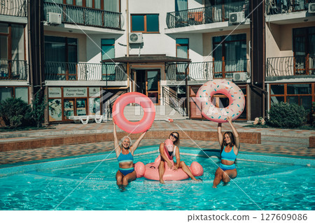 Three women are in a pool, holding pink inflatable donuts. Scene is fun and playful. Three women are in a pool, holding pink inflatable donuts. Scene is fun and playful. 127609086
