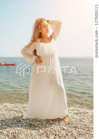 woman white dress stands on a beach, looking out at the ocean. She is in a contemplative or reflective mood, as she raises her hands above her head. Concept of peace and tranquility. 127609111