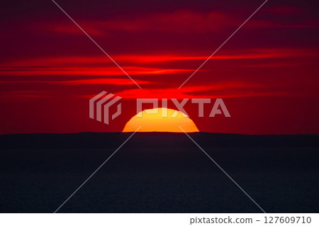 Red sunset over the sea horizon with the sun half-hidden behind a dark silhouette of distant hills. 127609710