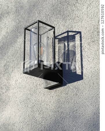 Modern outdoor wall light with a square design mounted on a textured wall. The metal frame encases a clear glass bulb, casting intricate shadows and creating a contemporary look 127610592