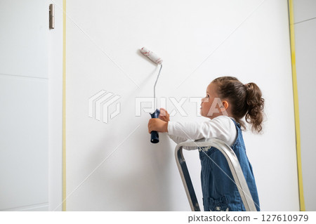 Child with curly hair painting a white wall in the apartment, cute girl with roller, standing on a ladder in denim overalls and a white sweater.  127610979