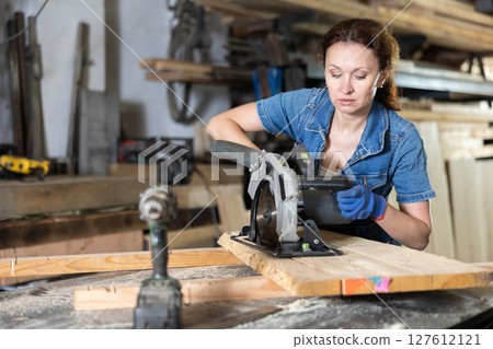 Female employee of workshop cuts wooden board into narrow parts using circular saw Female employee of workshop cuts wooden board into narrow parts using circular saw 127612121