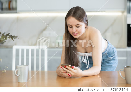 Interested girl standing in home kitchen, browsing through websites or chatting on social media on smartphone while leaning on table Interested girl standing in home kitchen, browsing through websites or chatting on social media on smartphone while leaning on table 127612124