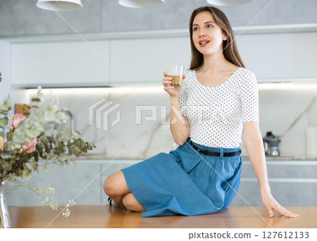 Portrait of cheerful contented woman with cup of cocoa in her hands in modern kitchen 127612133