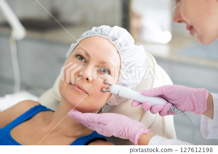 Woman getting facial radio wave lifting skin tightening procedure in salon. RF-lifting device treatment of acne scars, cellulite, skin rejuvenation, Concept of professional cosmetology and Woman getting facial radio wave lifting skin tightening procedure in salon. RF-lifting device treatment of acne scars, cellulite, skin rejuvenation, Concept of professional cosmetology and 127612198