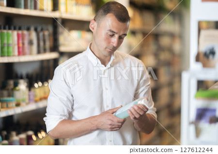 Thoughtful young man purchaser choosing cosmetic product in large supermarket 127612244