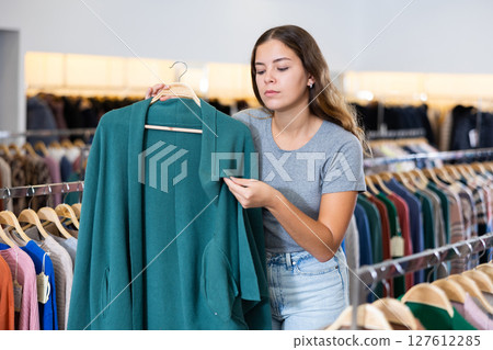 Serious young woman holding a turquoise cardigan on a rack while choosing clothes in a boutique Serious young woman holding a turquoise cardigan on a rack while choosing clothes in a boutique 127612285