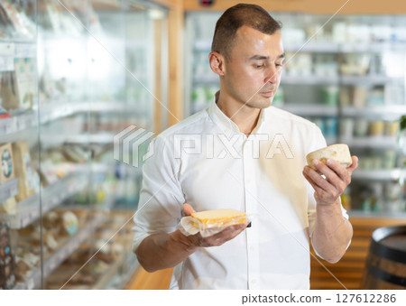 Attentive young man purchaser choosing cheese in large supermarket Attentive young man purchaser choosing cheese in large supermarket 127612286