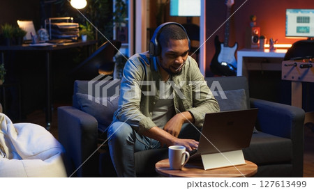 Focused black guy teleworking on laptop in a living room feels happy with task completion, achieving success. Remote worker balancing comfort and work efficiency in a flexible workspace. Camera B. Focused black guy teleworking on laptop in a living room feels happy with task completion, achieving success. Remote worker balancing comfort and work efficiency in a flexible workspace. Camera B. 127613499