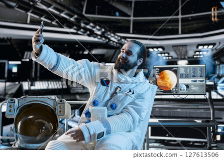Astronaut wearing spacesuit inside futuristic spacecraft doing selfie with smartphone. Smiling spaceman wearing equipment doing photos with mobile phone during space travel operations 127613506